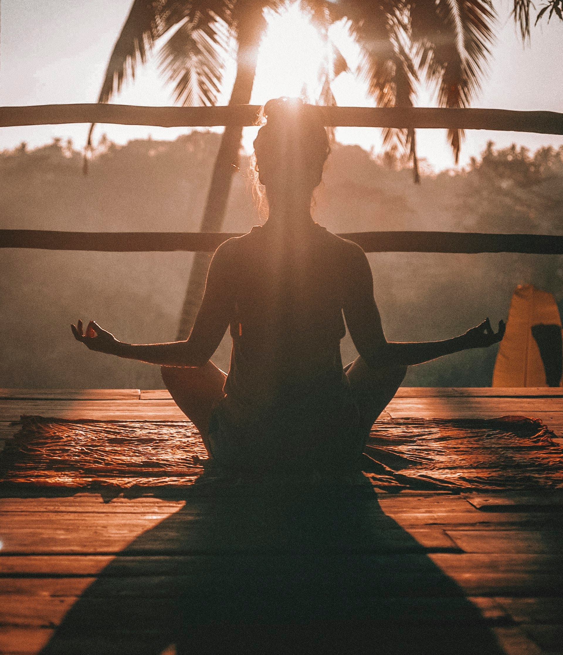 Yoga instructor in lotus meditation pose at golden sunrise on a wooden deck with palm trees